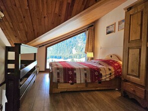Een slaapkamer met een houten plafond, een bed met een bedovertrek met patroon, een raam met uitzicht op een bos en een houten bank.