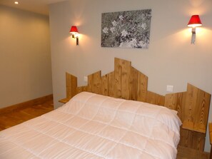 A bedroom with a wooden headboard, a bed with a white quilt, two wall-mounted lamps, and a framed picture on the wall. - Charming apartment - 7 guests (Pralognan-la-Vanoise)