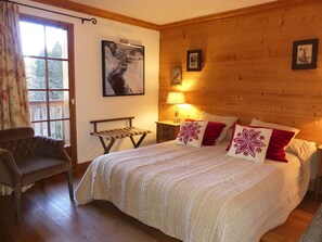 A cozy bedroom with a wooden headboard, a large bed with white bedding and red pillows, a bench, and a window with curtains. - Spacious apartment with balcony - 8 guests (Pralognan-la-Vanoise)