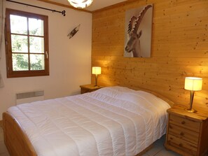 A bedroom with a large bed, wooden headboard, two bedside tables with lamps, a window with curtains, and a wall-mounted painting of a deer. - Comfortable apartment near the center - 4 guests (Pralognan-la-Vanoise)