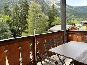 Eine Holzterrasse mit Tisch und Stühlen, die auf eine gebirgige Landschaft mit Bäumen und Gebäuden blickt.