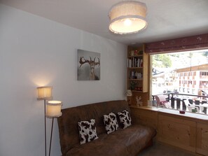 Ein gemütliches Wohnzimmer mit einem braunen Sofa, zwei Kissen mit Kuhmuster, einem an der Wand hängenden Hirschgemälde, einem Bücherregal und einem Fenster mit Blick auf Gebäude.