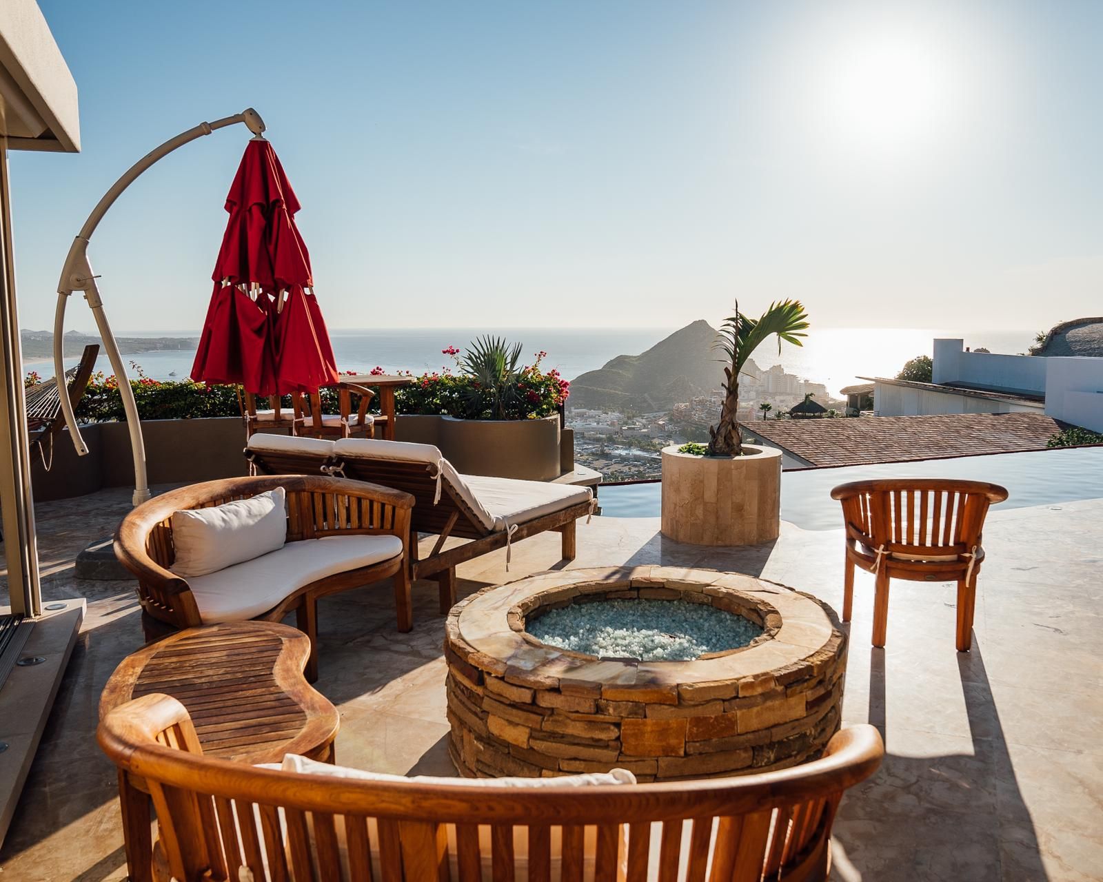 A sunlit outdoor seating area with a stone fire pit, lounge chairs, and a red umbrella.