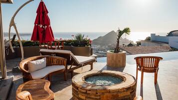 A sunlit outdoor seating area with a stone fire pit, lounge chairs, and a red umbrella.