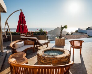 A sunlit outdoor seating area with a stone fire pit, lounge chairs, and a red umbrella. - Pedregal Villa with 75-ft Infiniti Pool + Panoramic Views (Cabo San Lucas)