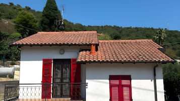 Pequeño edificio blanco con techo de tejas rojas y contraventanas rojas, rodeado de hierba seca y una colina con árboles.