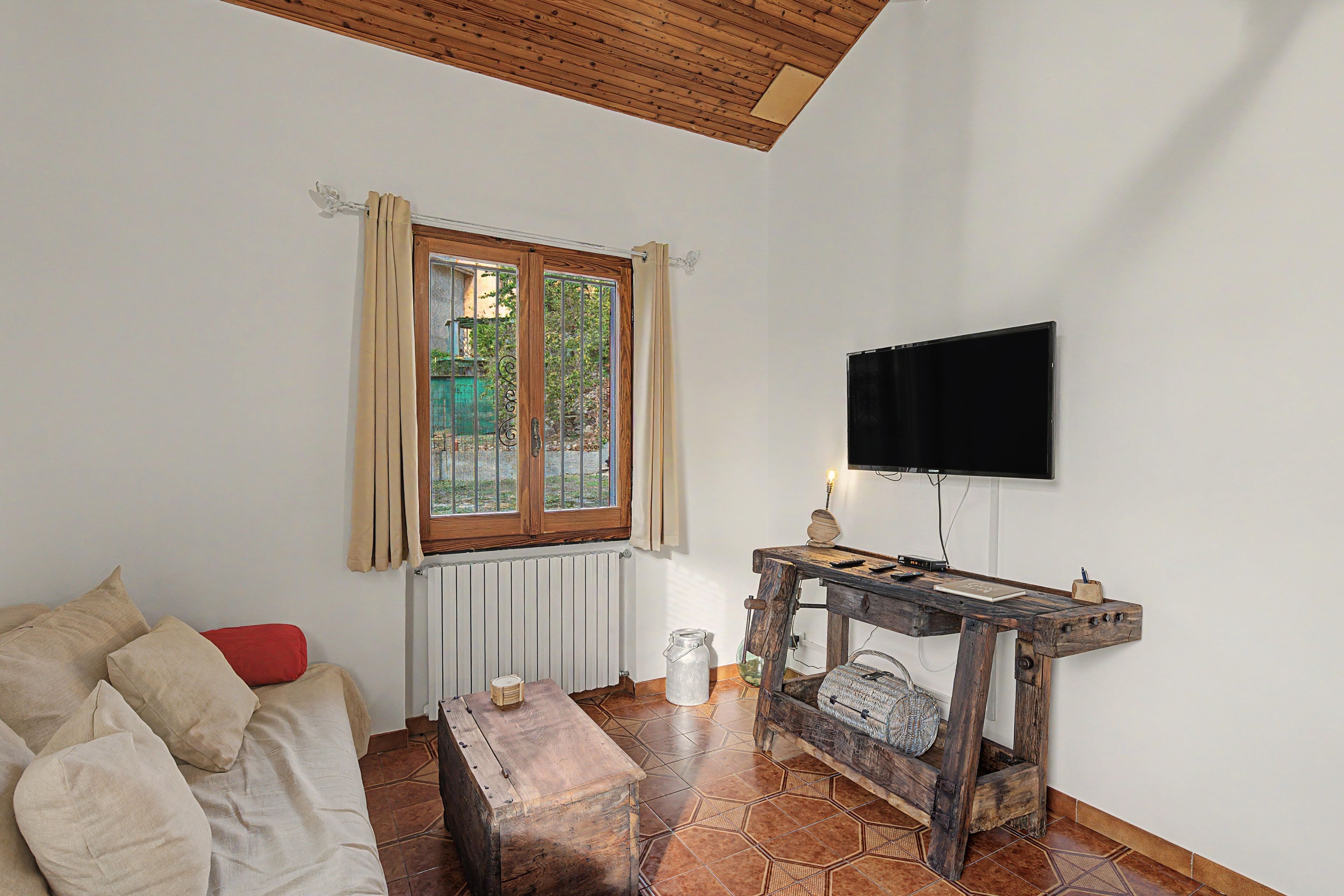 A room with a wooden ceiling, a window with curtains, a television mounted on the wall, a wooden table with various items, and a sofa with cushions.