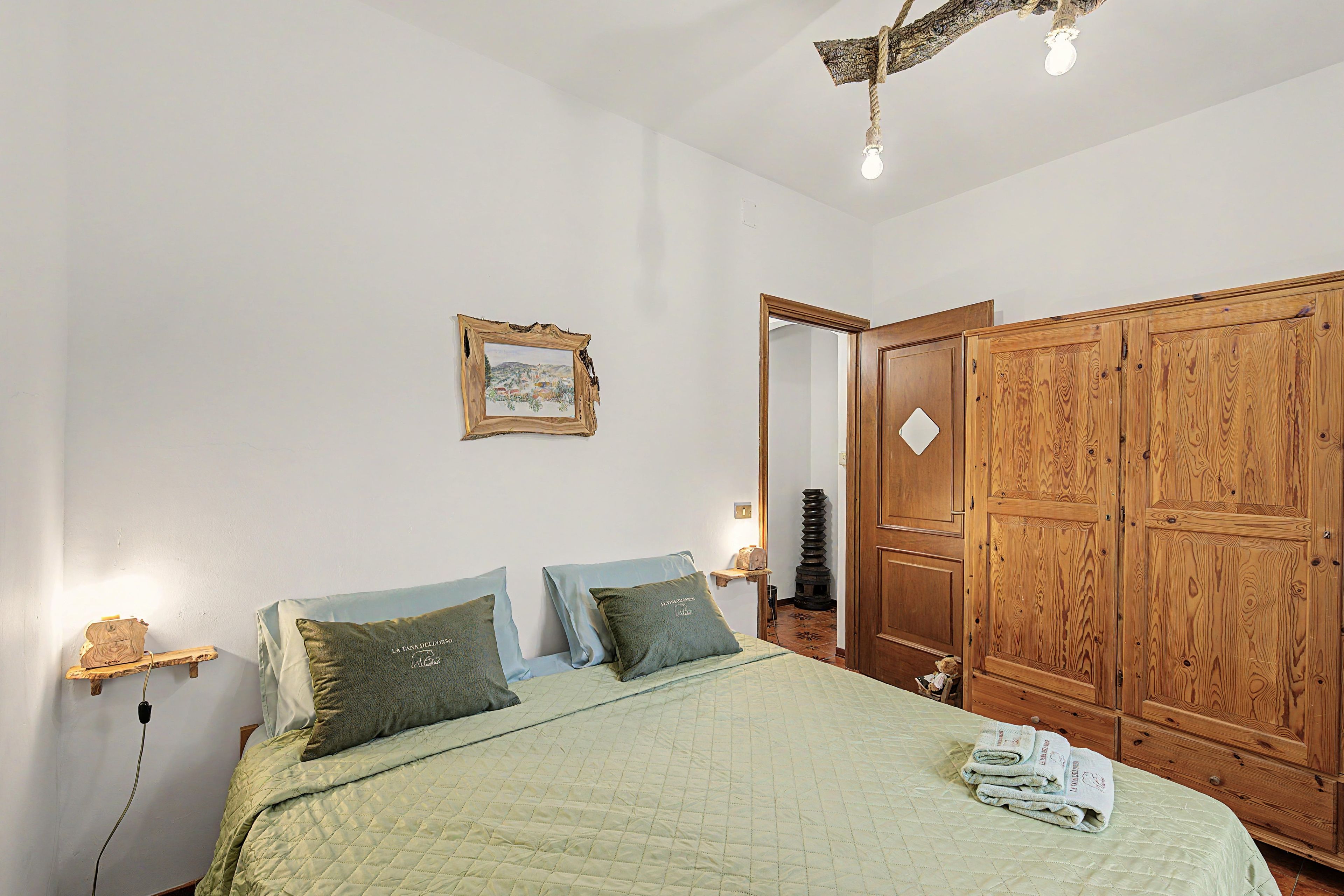 A bedroom with a bed, pillows, a wooden wardrobe, a framed picture on the wall, and a ceiling light fixture.