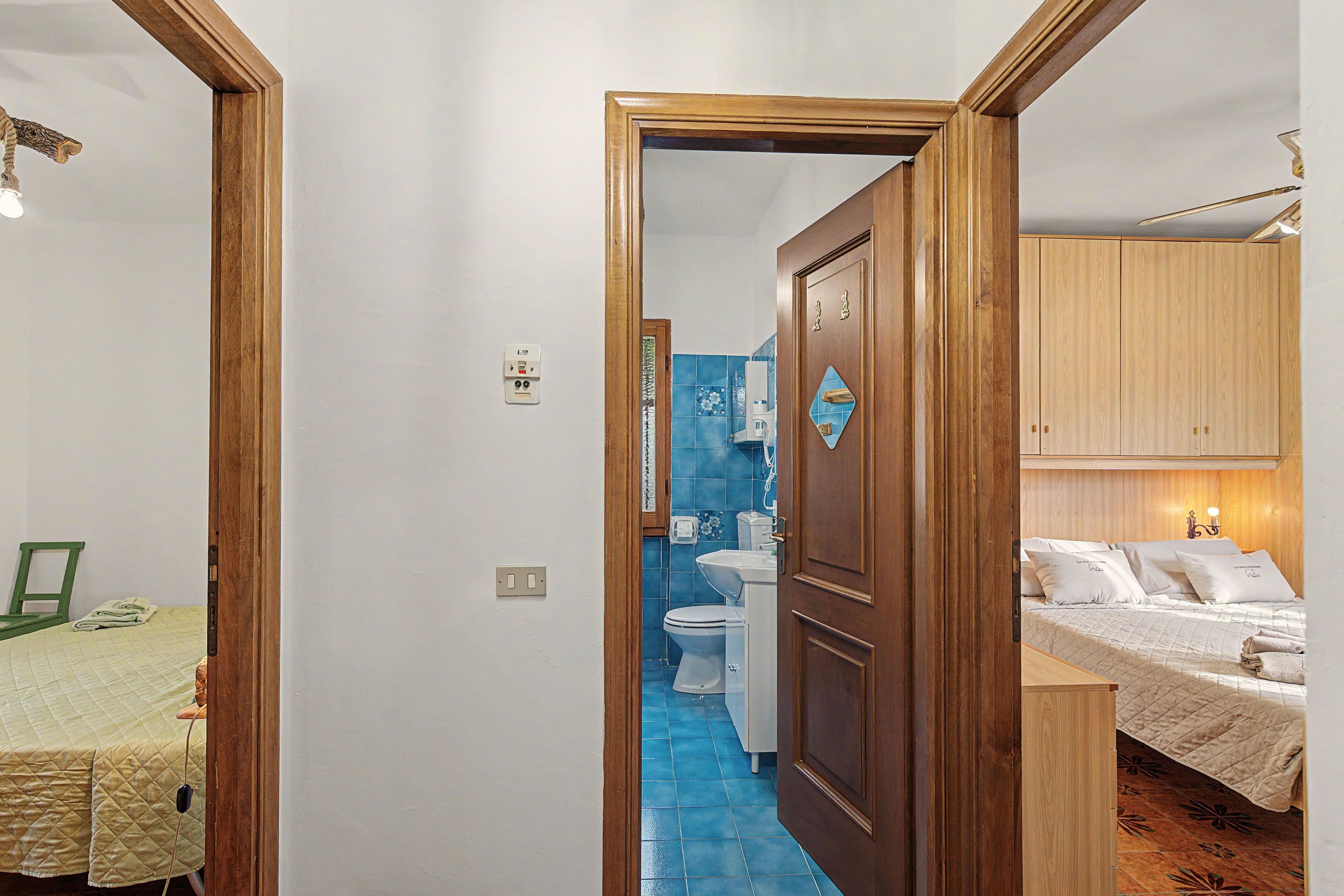 Un couloir étroit mène à une salle de bain équipée de toilettes et d’un lavabo, ainsi qu’à une chambre avec un lit recouvert d’une courtepointe et une armoire en bois.