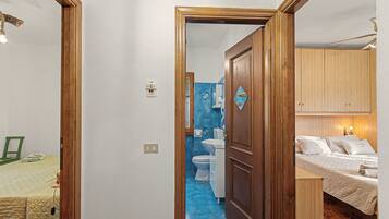 Un couloir étroit mène à une salle de bain équipée de toilettes et d’un lavabo, ainsi qu’à une chambre avec un lit recouvert d’une courtepointe et une armoire en bois.