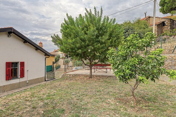 A residential area with a house featuring a red window shutter, a tree, and a stone wall in the background.