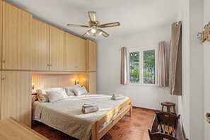 A bedroom with a bed, wooden wardrobe, ceiling fan, window with curtains, and a small wooden stool.