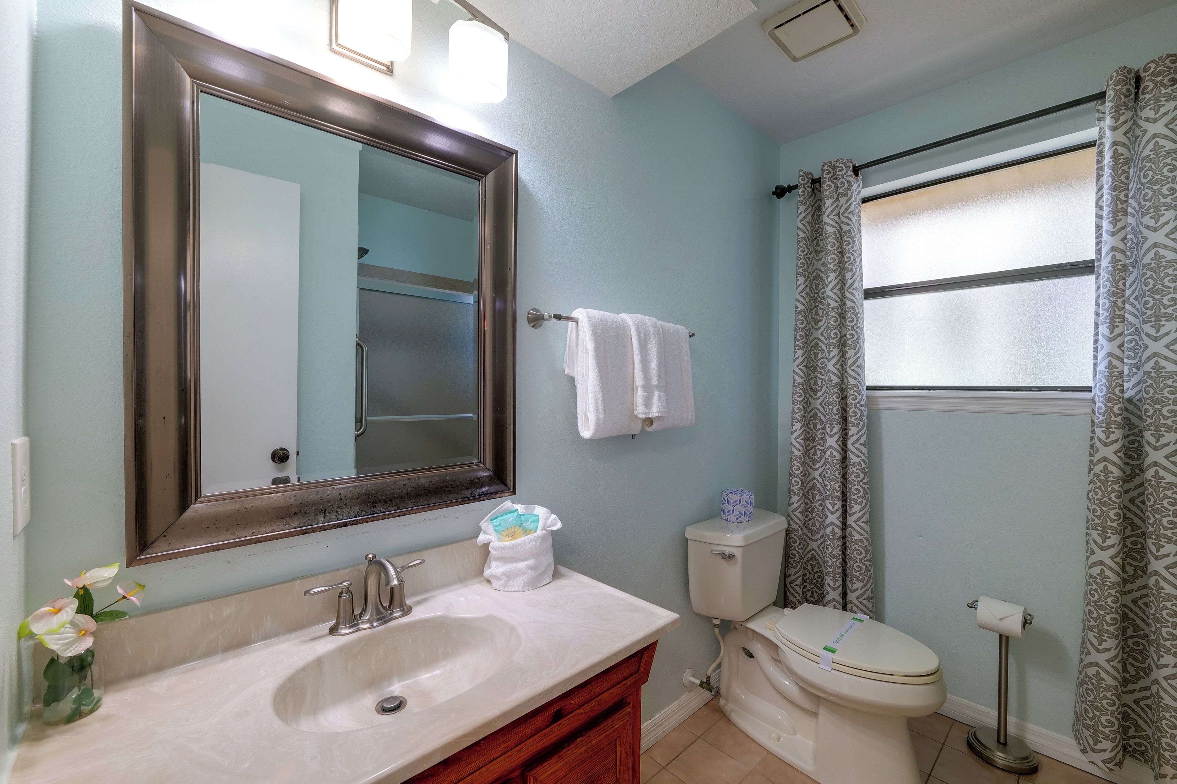 A bathroom with a sink, mirror, toilet, and window with patterned curtains.