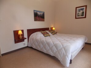 Cama bem arrumada com colcha branca, duas luminárias de cabeceira e dois quadros na parede.