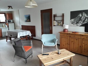 A living room with a dining area, a light blue armchair, a wooden table, and a bookshelf with books. - Spacious apartment for families - 6 guests (Pralognan-la-Vanoise)