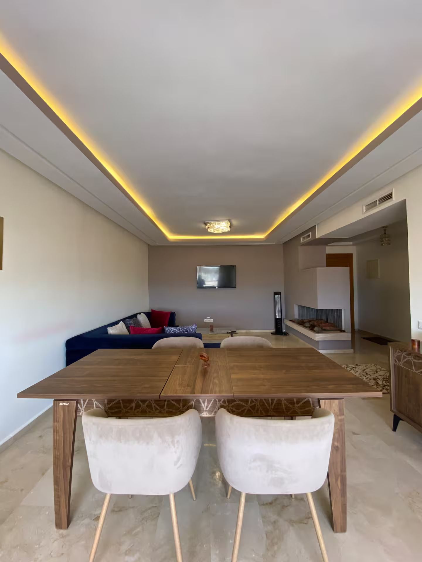 Una sala de estar moderna con una mesa de comedor de madera, dos sillas blancas, un sofá azul y un televisor de pantalla plana montado en la pared.