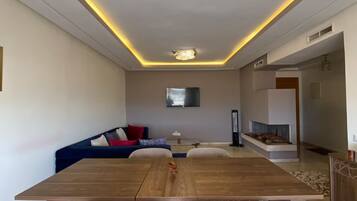 Una sala de estar moderna con una mesa de comedor de madera, dos sillas blancas, un sofá azul y un televisor de pantalla plana montado en la pared.