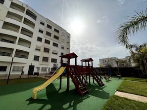 Un parque infantil con un tobogán y estructuras para escalar, rodeado por un edificio con varios balcones.