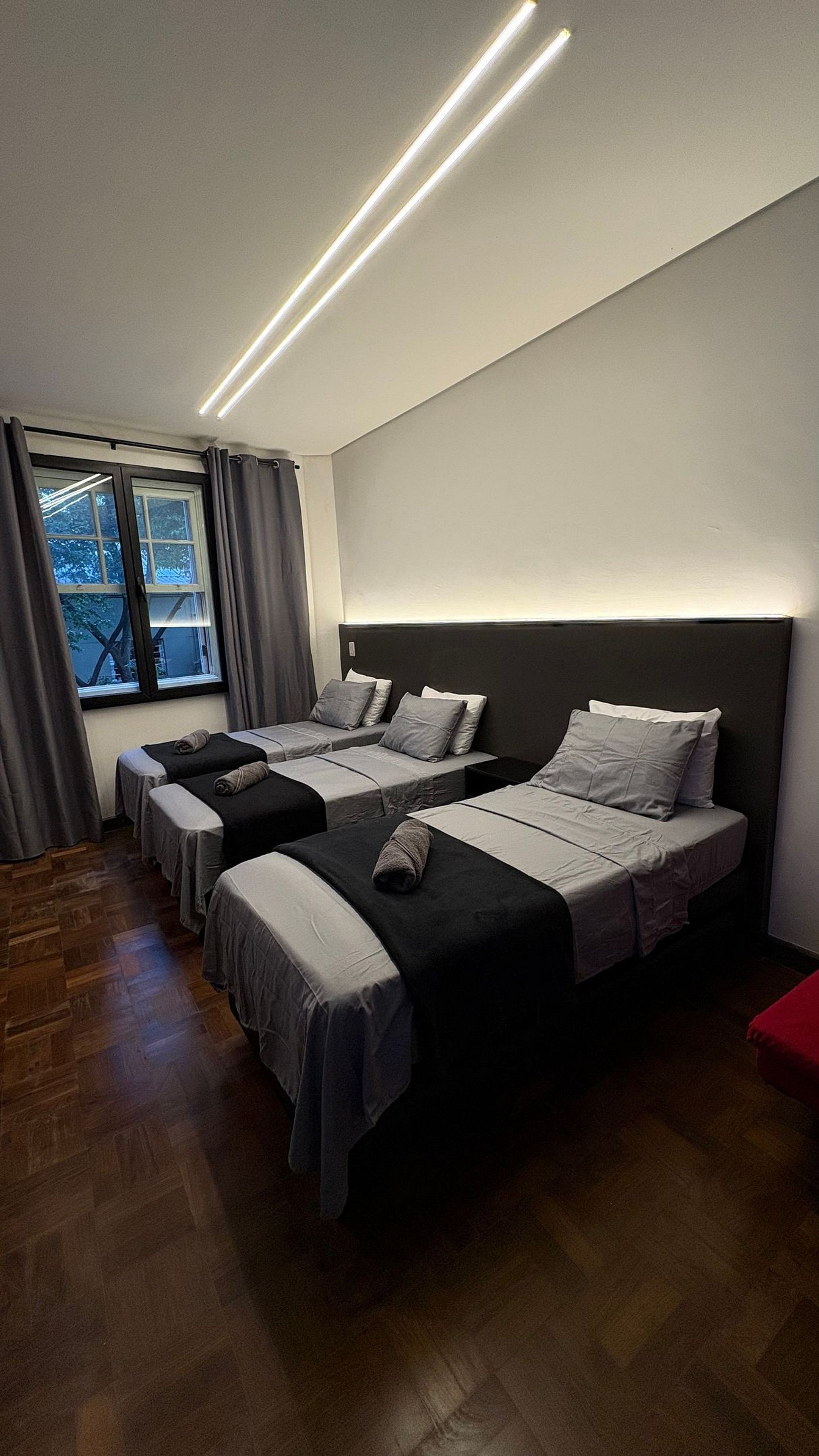 Ein Hotelzimmer mit drei nebeneinanderstehenden Betten, einem großen Fenster mit Vorhängen und einer modernen Deckenleuchte.