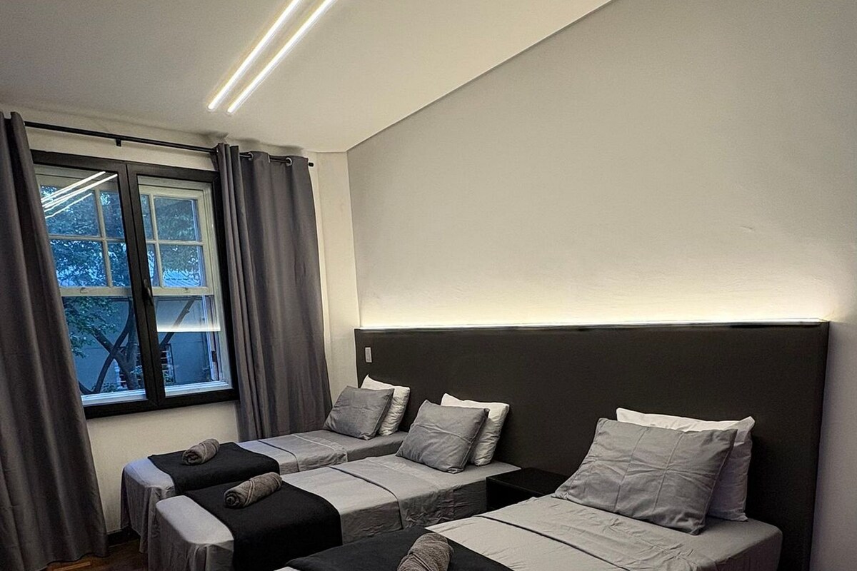 Une chambre d’hôtel avec trois lits disposés en rangée, une grande fenêtre avec des rideaux et un luminaire moderne au plafond.