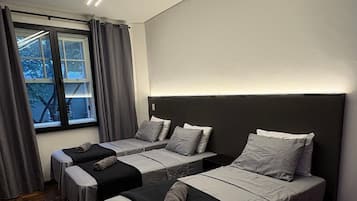 Ein Hotelzimmer mit drei nebeneinanderstehenden Betten, einem großen Fenster mit Vorhängen und einer modernen Deckenleuchte.