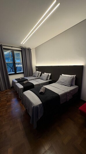 Une chambre d’hôtel avec trois lits disposés en rangée, une grande fenêtre avec des rideaux et un luminaire moderne au plafond.