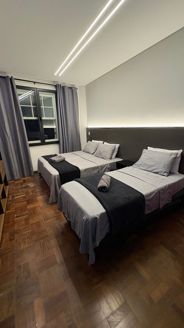 Habitación de hotel con dos camas, piso de madera, ventana con cortinas y una lámpara de techo moderna.