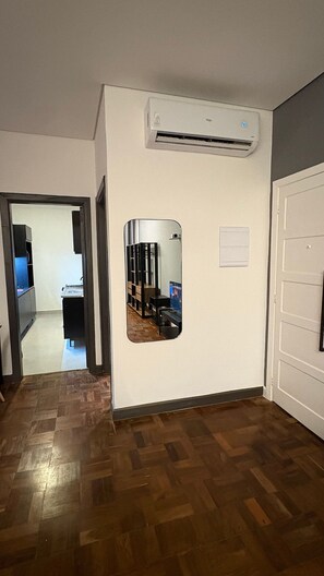 Una habitación con piso de madera, pared blanca con aire acondicionado instalado, una puerta y un pasillo que conduce a otra habitación.