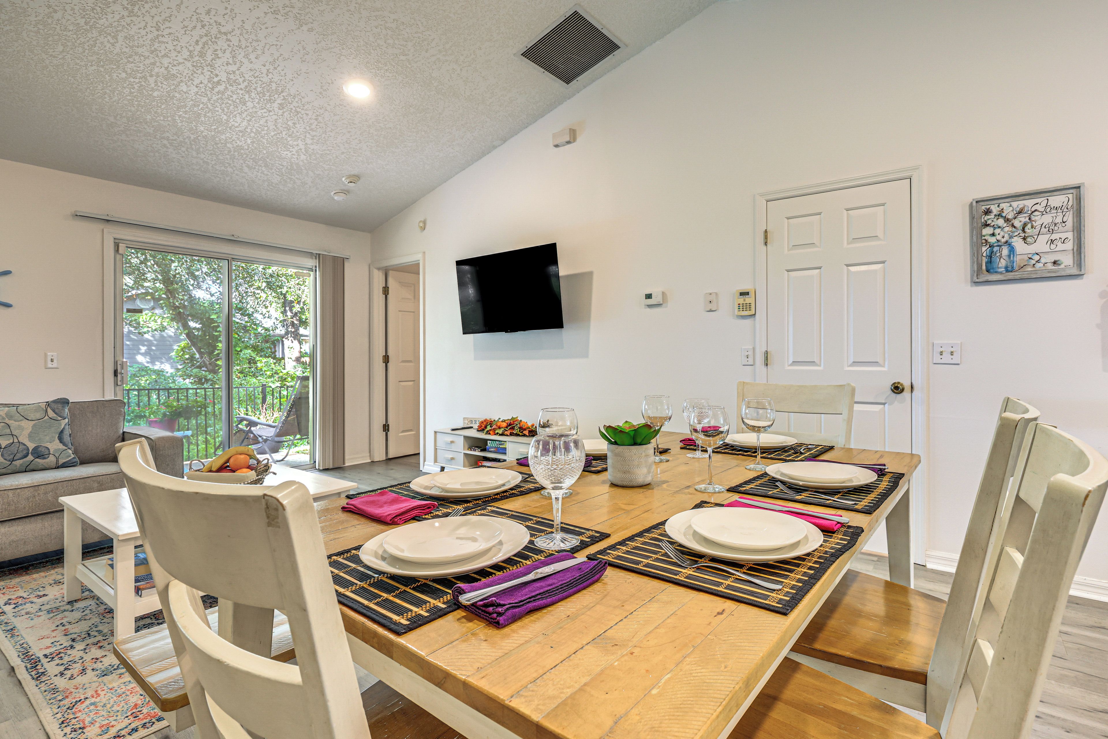 Uma sala de jantar com mesa de madeira posta para quatro, cadeiras brancas e televisão na parede.