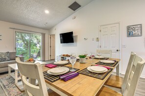 Uma sala de jantar com mesa de madeira posta para quatro, cadeiras brancas e televisão na parede.