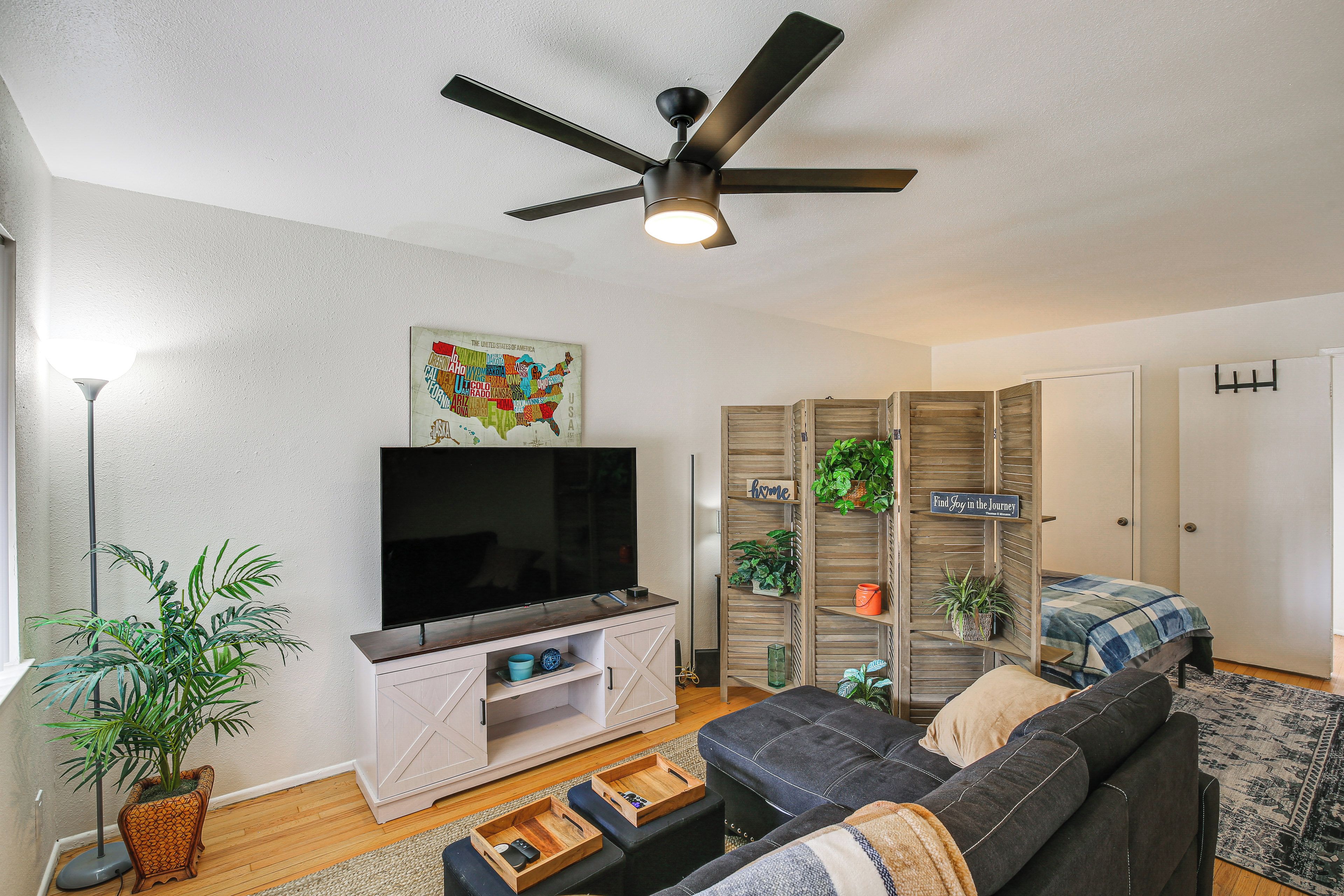 A living room with a flat-screen TV, a ceiling fan, a map on the wall, and a bookshelf with plants.