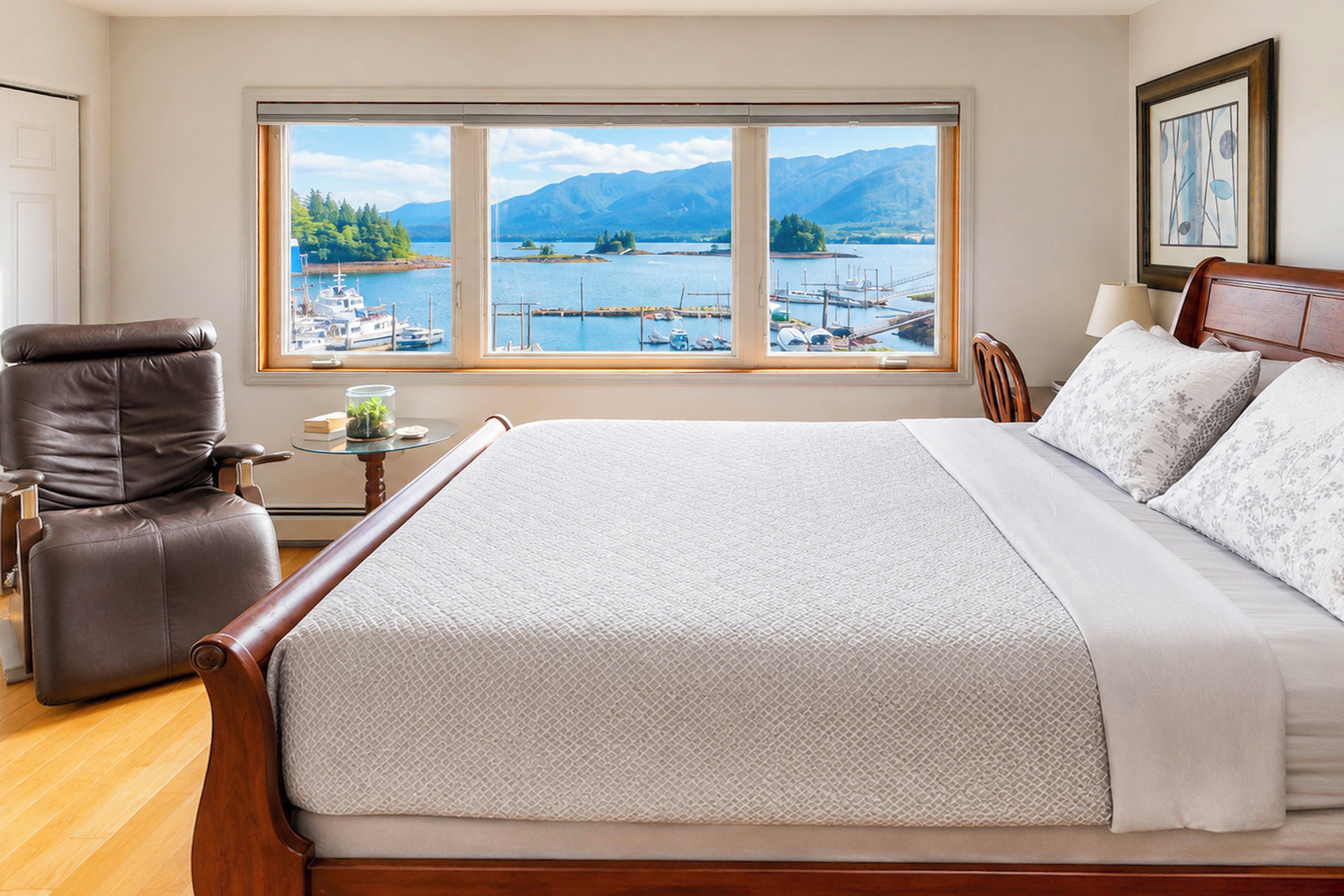 Une chambre avec un grand lit, une chaise, une petite table sur laquelle se trouve un récipient en verre, et une vue sur une marina et des montagnes, visible à travers une grande fenêtre.
