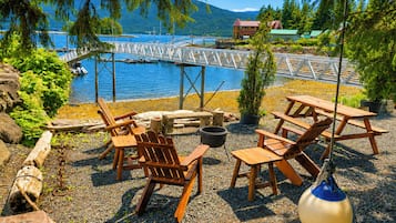 A lakeside outdoor seating area with wooden chairs and tables, a grill, and a view of the lake and mountains.