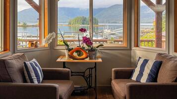 A cozy room with two brown sofas, a small table with potted plants, and a view of a marina and mountains through large windows.