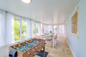 Una habitación con mesa de fútbol, una mesa de comedor con sillas y amplias ventanas con cortinas.