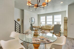Ein Essbereich mit Glastisch, weißen Stühlen, einer Kronleuchter und Blick auf ein Wohnzimmer mit Sofa und Fenstern.