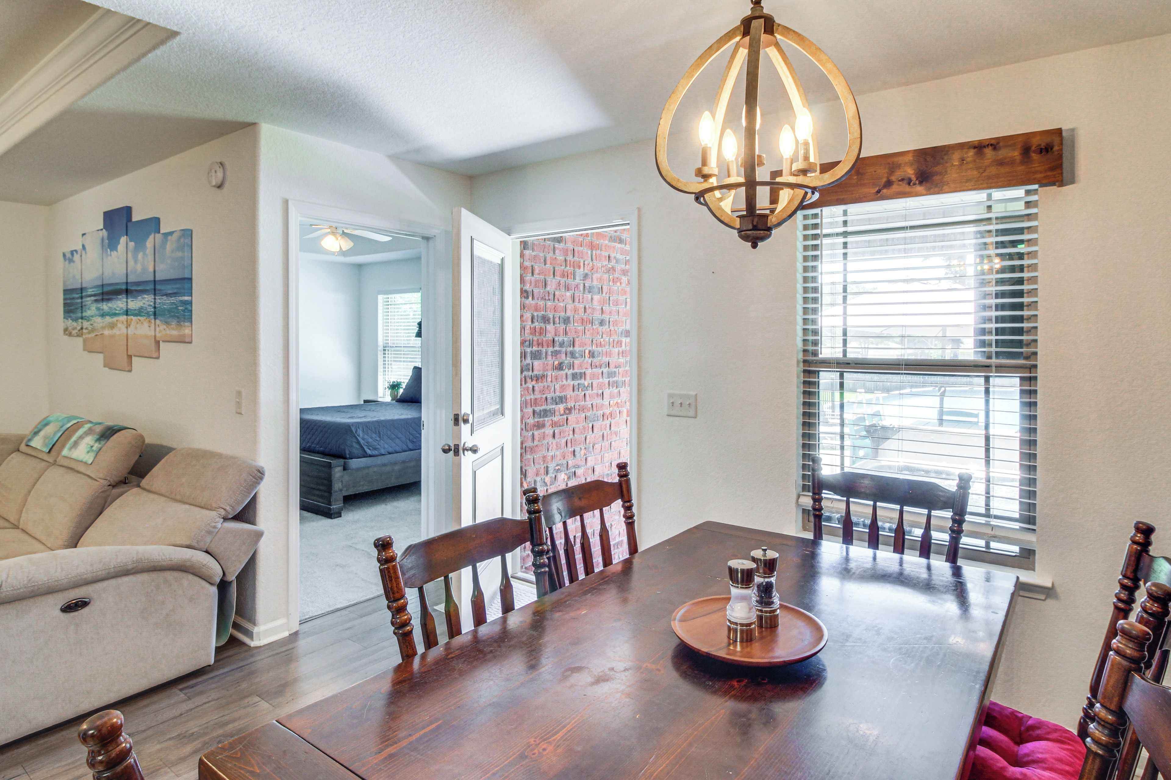 A dining area with a wooden table, chairs, and a chandelier. There is a brick wall and a window with blinds.