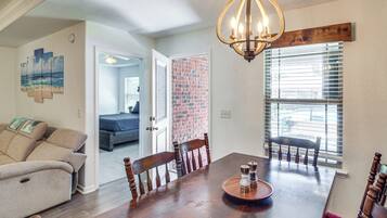 A dining area with a wooden table, chairs, and a chandelier. There is a brick wall and a window with blinds.
