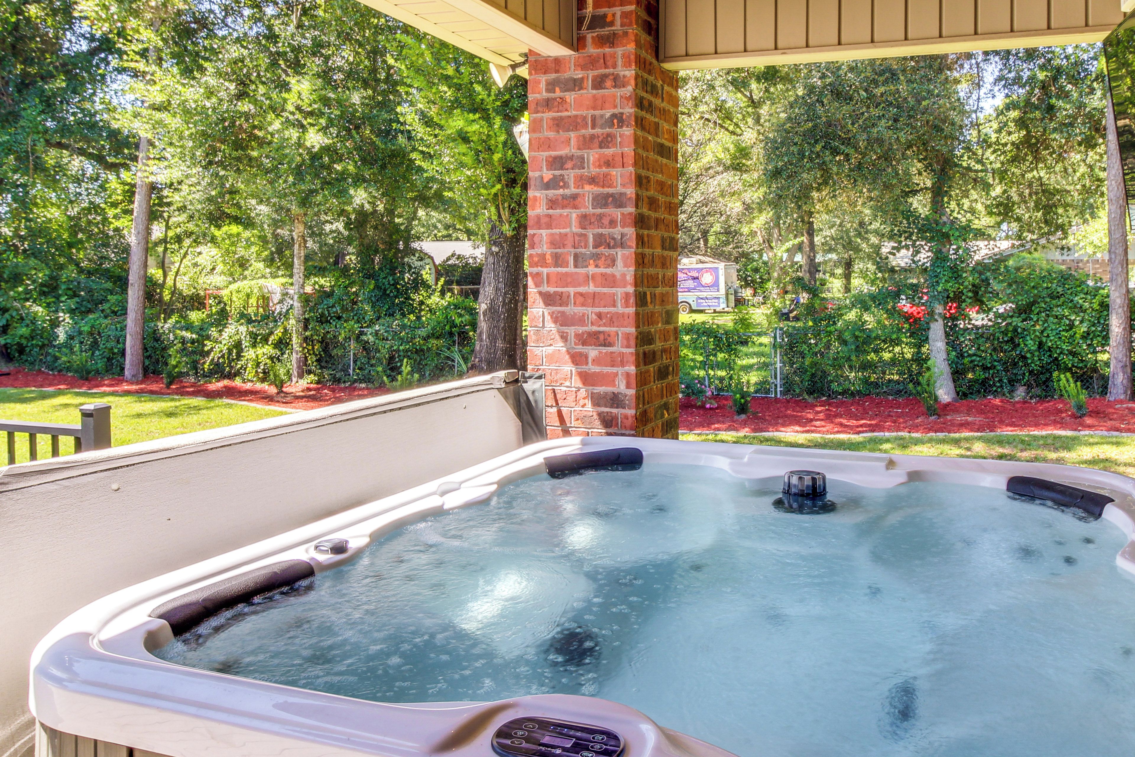 A hot tub in an outdoor setting with a brick pillar, surrounded by lush greenery and a covered area.