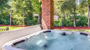 A hot tub in an outdoor setting with a brick pillar, surrounded by lush greenery and a covered area.