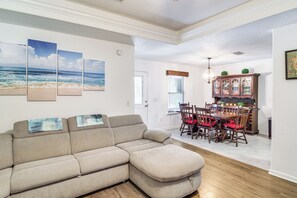 A living room with a beige sectional sofa, a dining area with wooden chairs and a table, and a kitchen visible in the background.