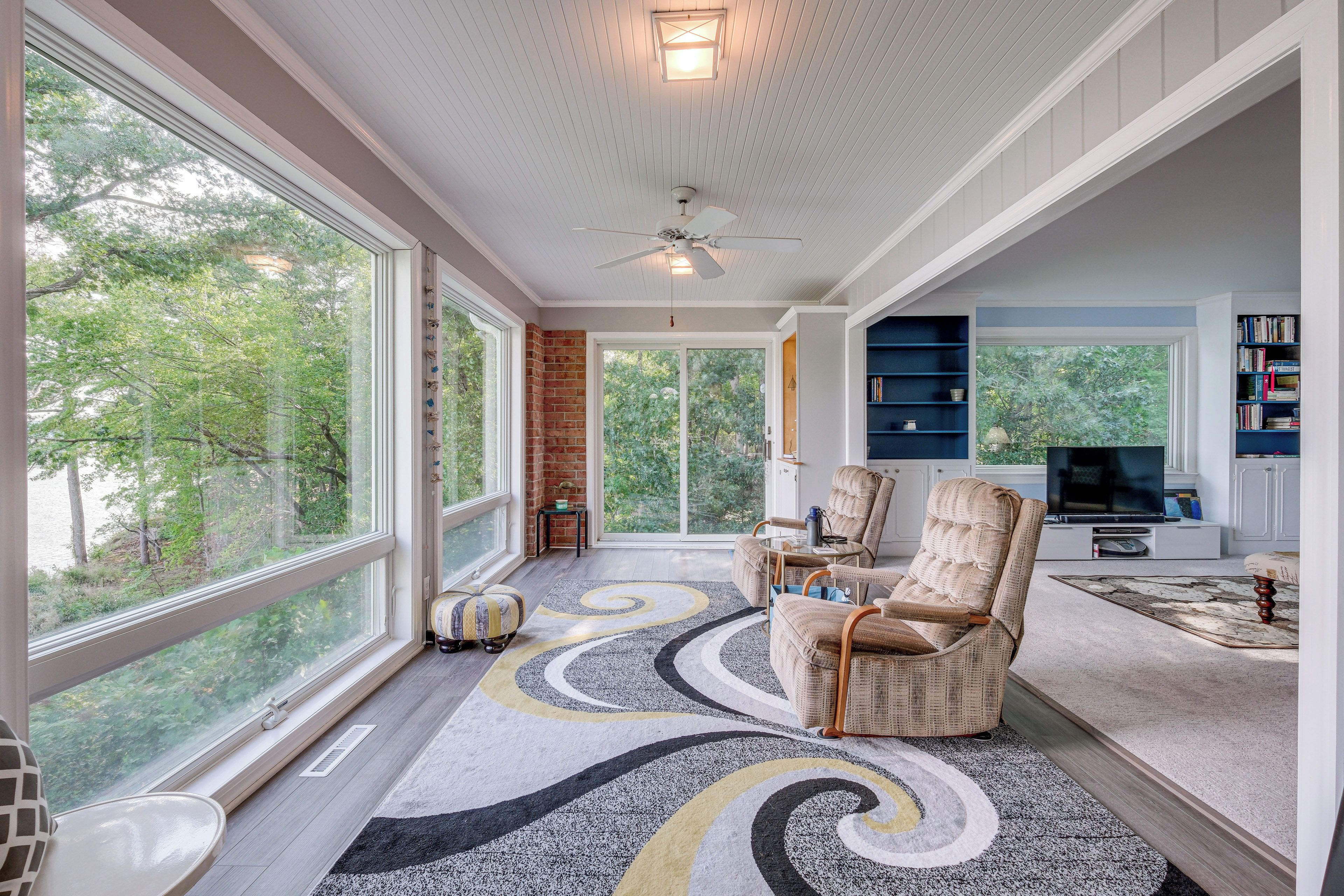 A spacious living room with large windows, a fireplace, a ceiling fan, and a television.