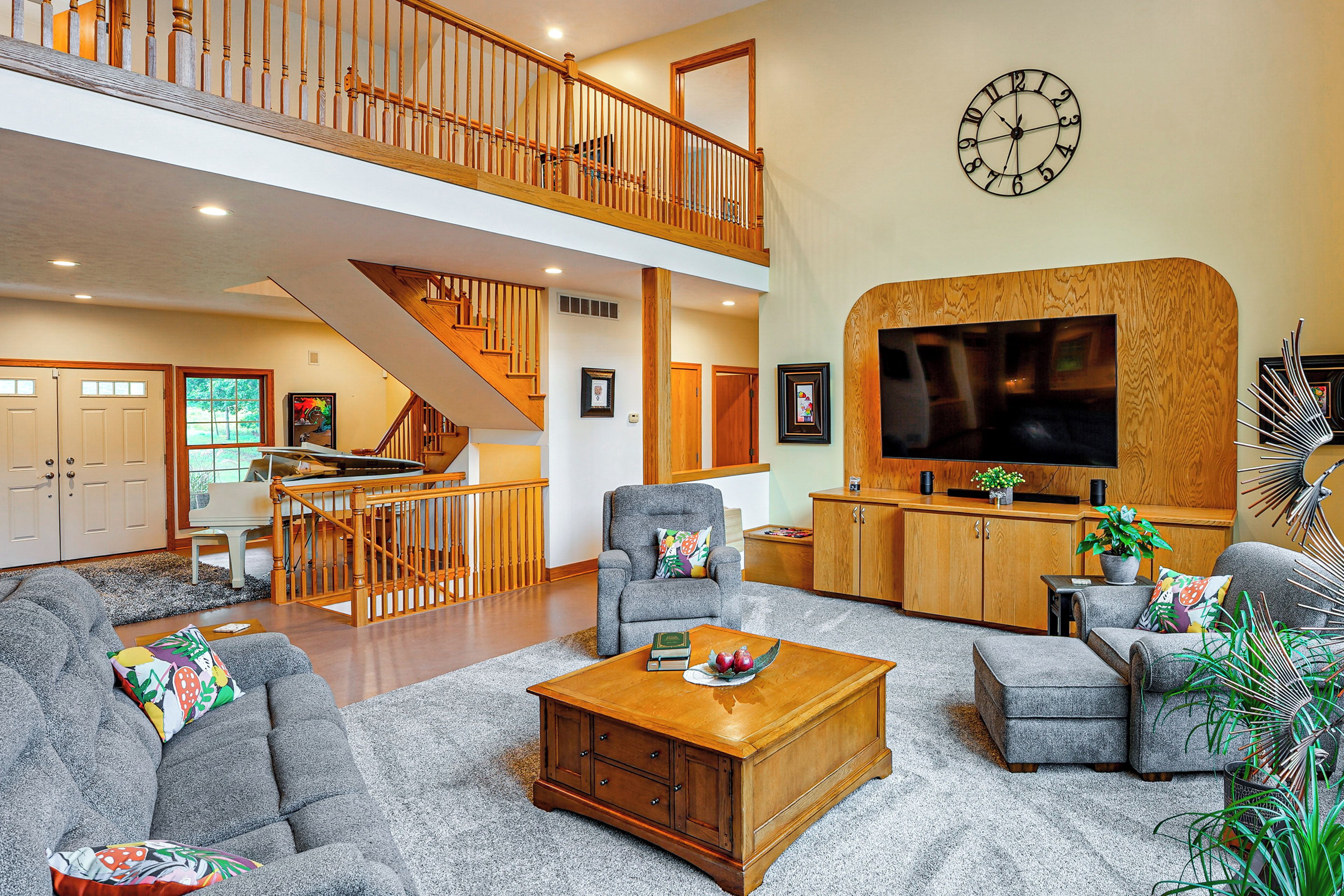 A spacious living room with a high ceiling, a staircase, and a large flat-screen TV mounted on a wooden wall.