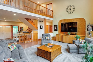A spacious living room with a high ceiling, a staircase, and a large flat-screen TV mounted on a wooden wall. - Spacious Executive Home on Over 11 Private Acres (Rockford)