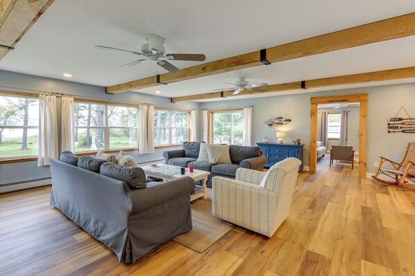 A spacious living room with a large gray sectional sofa, a smaller striped armchair, a coffee table, and a blue cabinet. There are two ceiling fans, large windows with curtains, and a wooden floor.