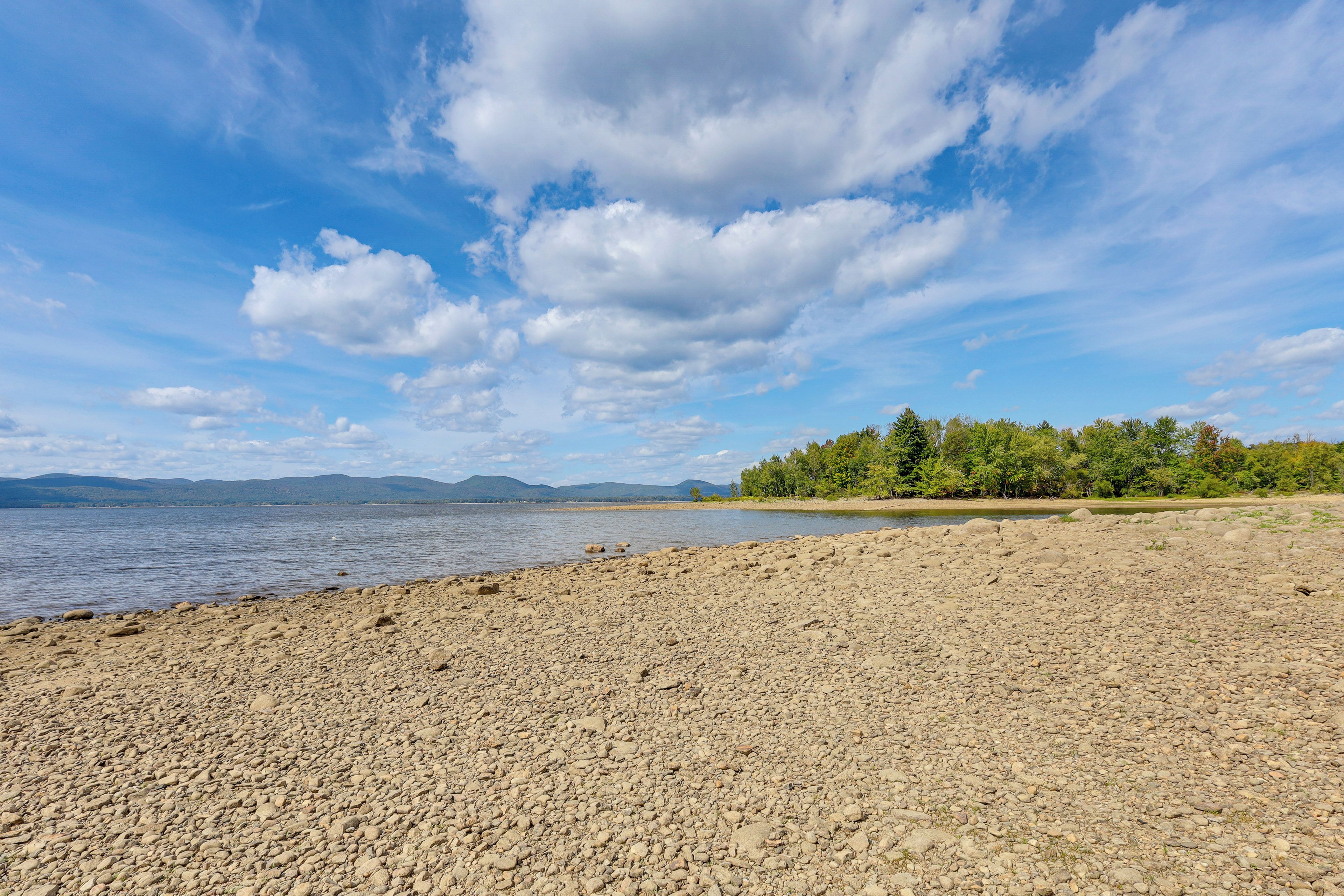 A pebbly beach with a calm lake, trees, and distant mountains under a partly cloudy sky.