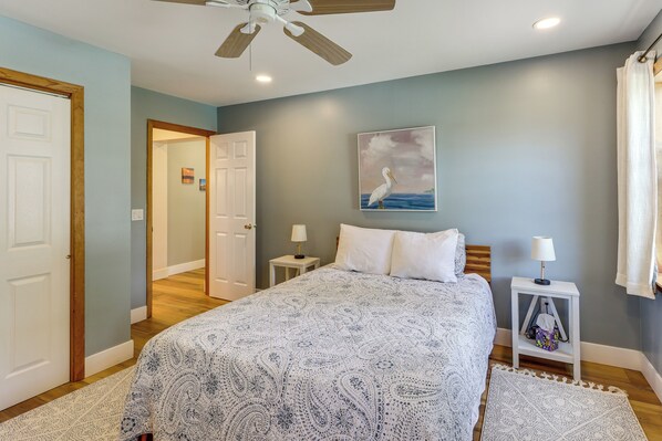A bedroom with a bed, two bedside tables, a ceiling fan, a painting, and a view of a hallway. - Lakefront Adirondack Escape! Fire Pit & Game Room (Broadalbin)