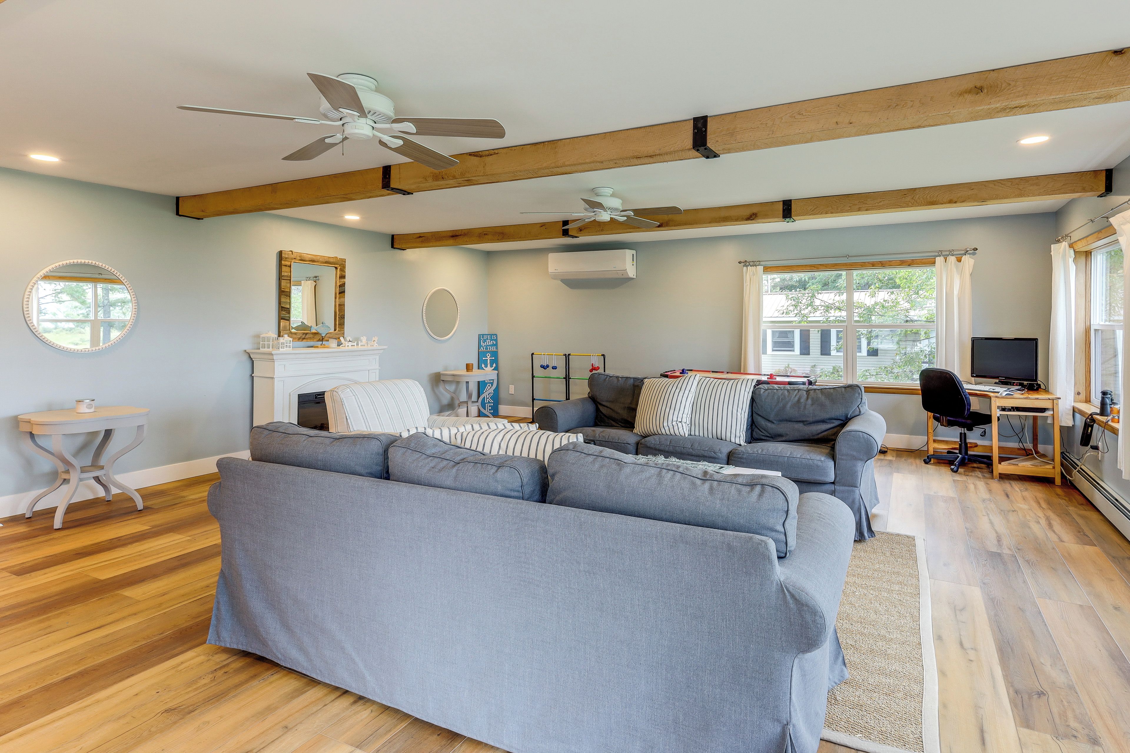 Ein geräumiges Wohnzimmer mit einem großen, grauen Ecksofa, einem Kamin, einem Deckenventilator und einem großen Fenster mit Vorhängen.