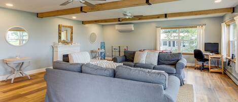 Ein geräumiges Wohnzimmer mit einem großen, grauen Ecksofa, einem Kamin, einem Deckenventilator und einem großen Fenster mit Vorhängen.