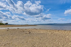A pebbly beach with a calm lake and distant mountains under a partly cloudy sky. - Lakefront Adirondack Escape! Fire Pit & Game Room (Broadalbin)